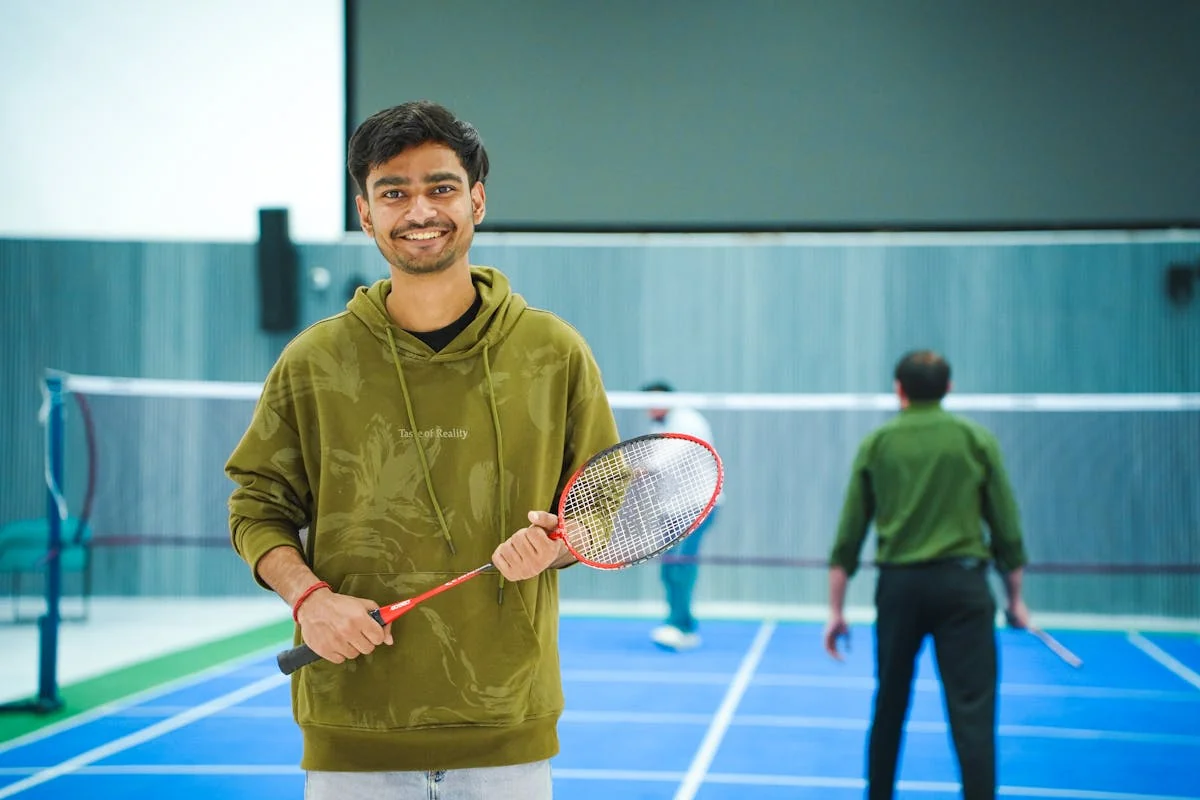 Young badminton player holding racket confidently at indoor court with other players practicing in background