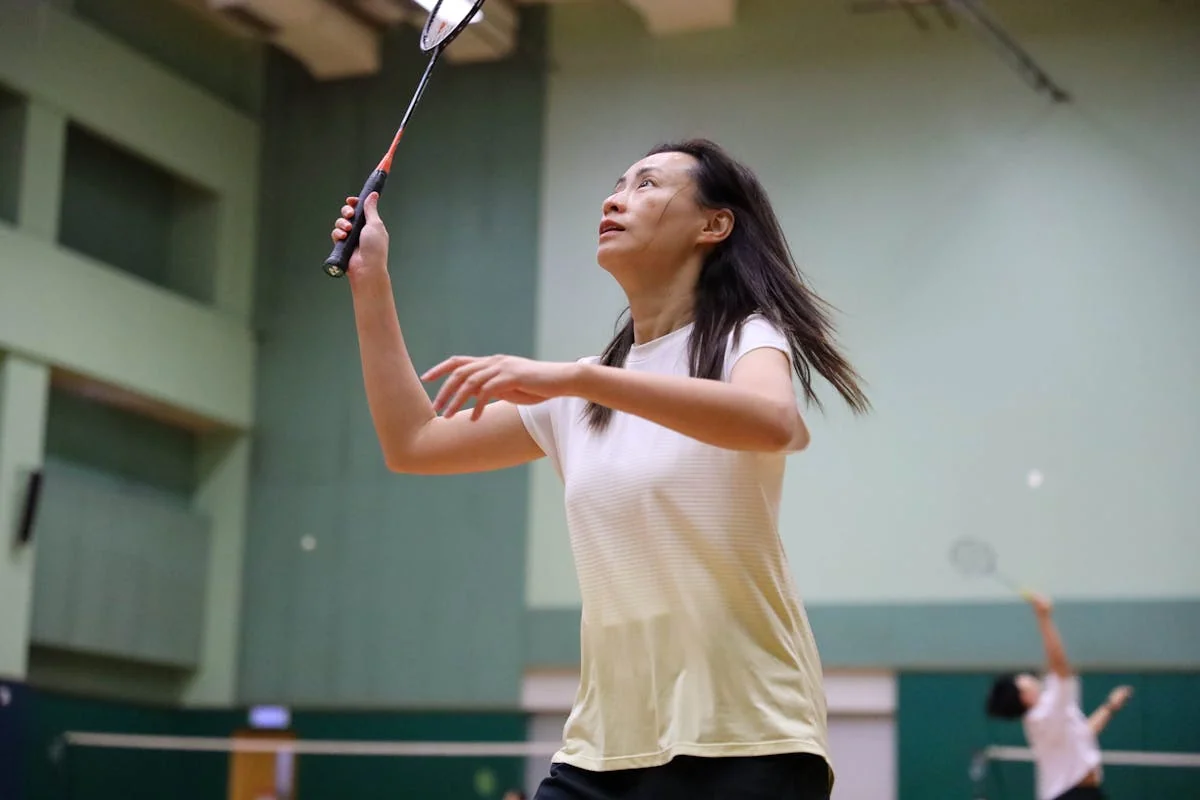 Female badminton player looking up at shuttlecock during indoor doubles match with second player visible in background