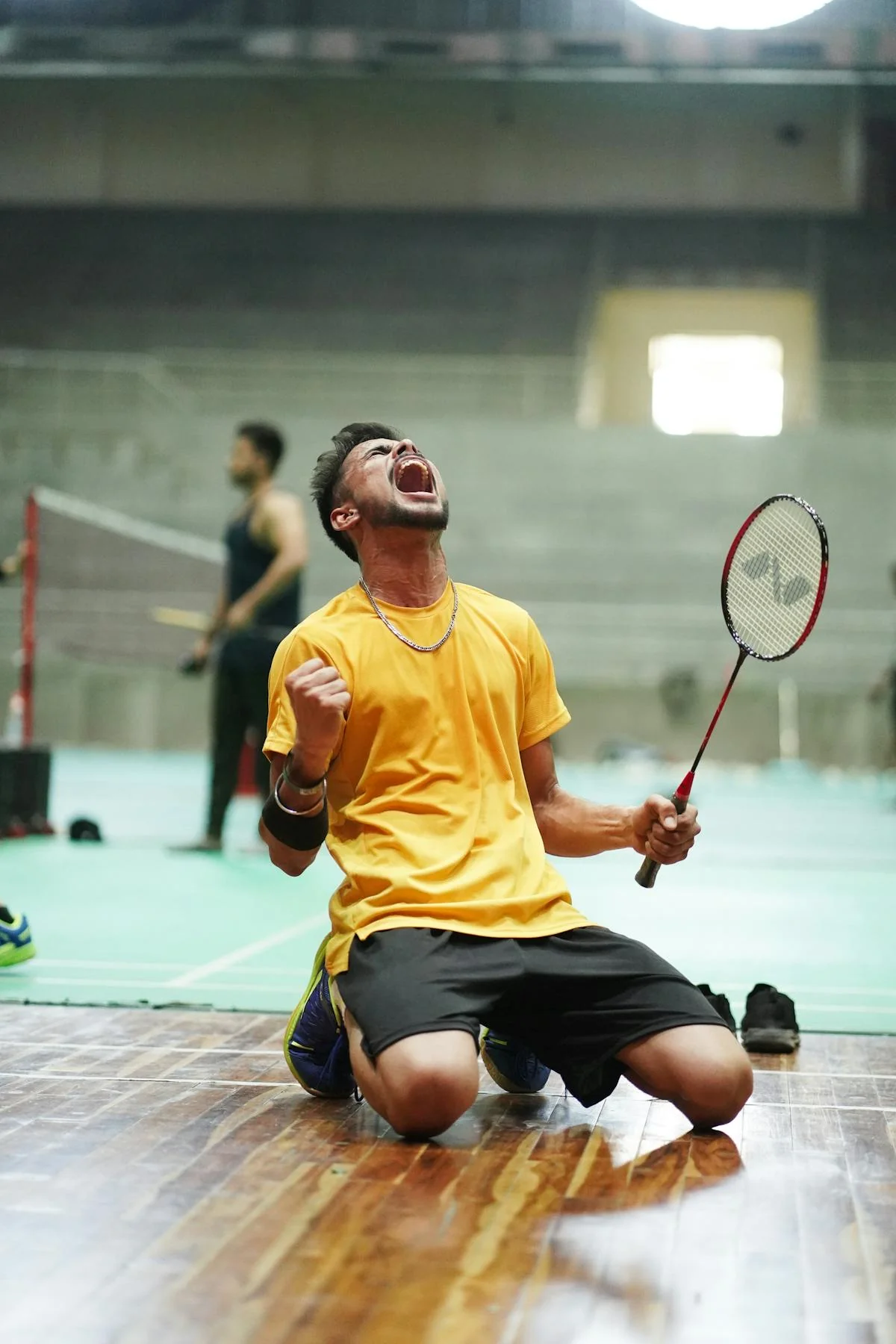 Male badminton player kneeling on indoor court celebrating a match victory with racket raised